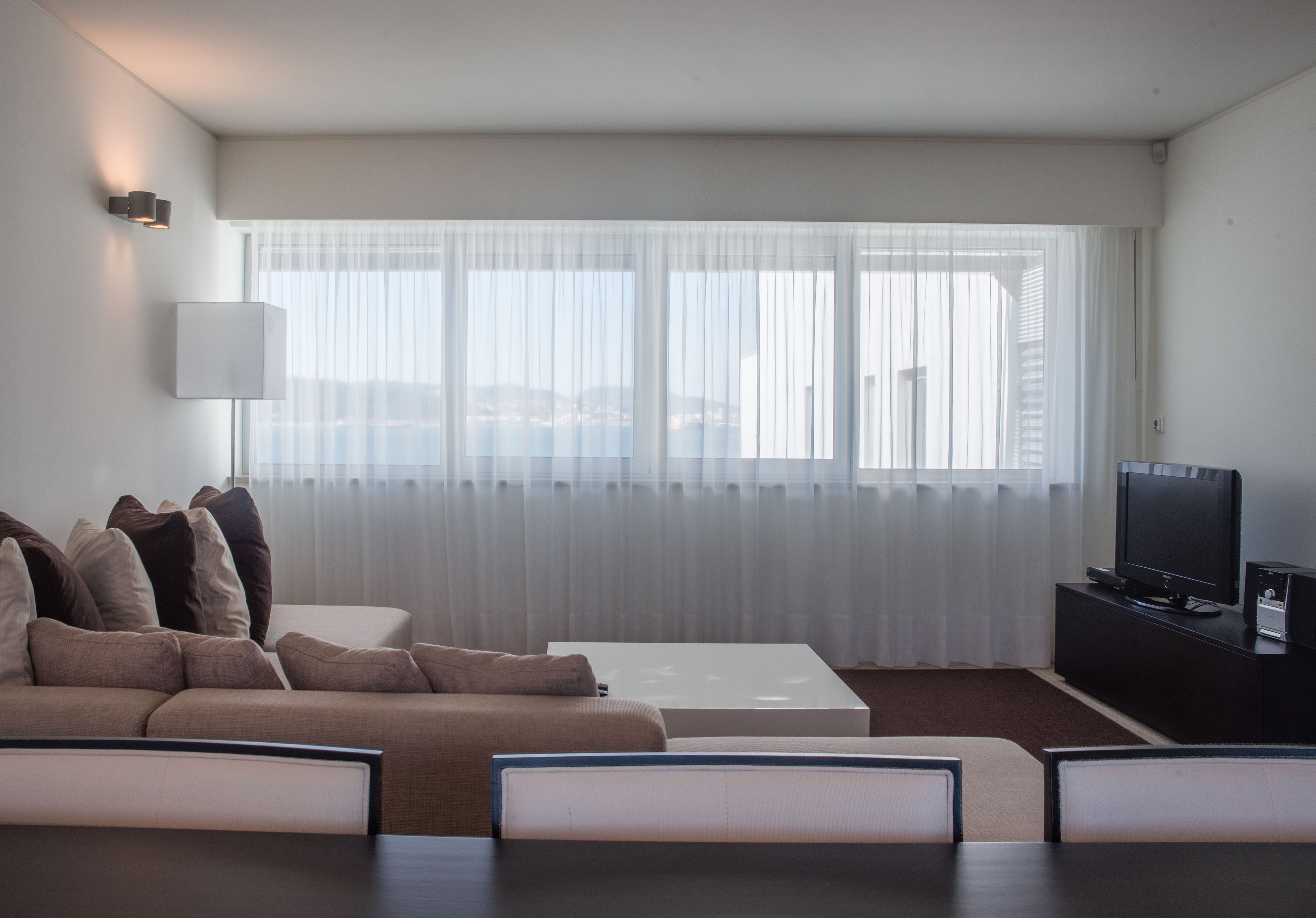 Uma sala de estar moderna com um sofá, uma mesa de centro, uma TV sobre um suporte preto e cortinas translúcidas nas janelas.
