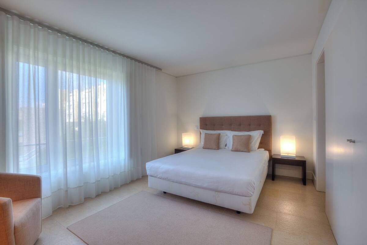 Un dormitorio moderno con una cama grande, dos mesitas de noche con lámparas, un cabecero de madera y cortinas translúcidas.