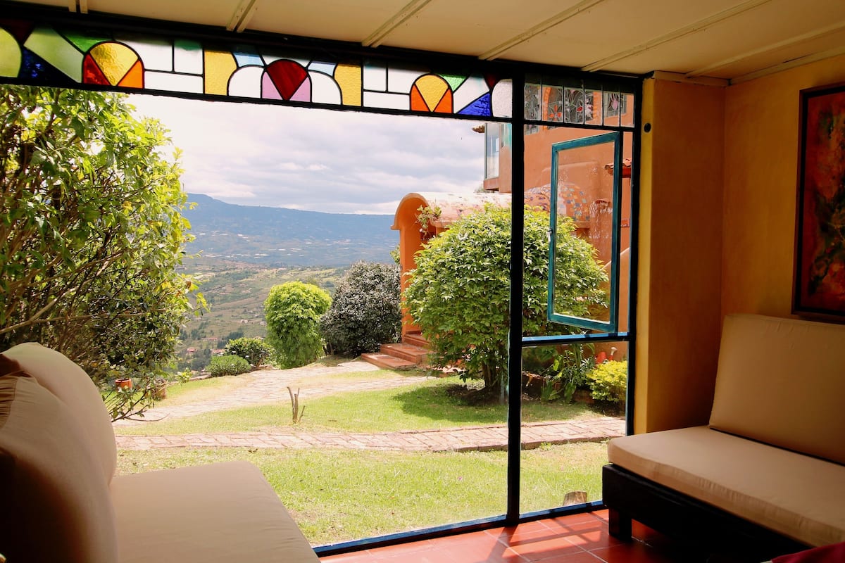 Una habitación con un ventanal amplio que ofrece vistas a un jardín frondoso y colinas distantes.