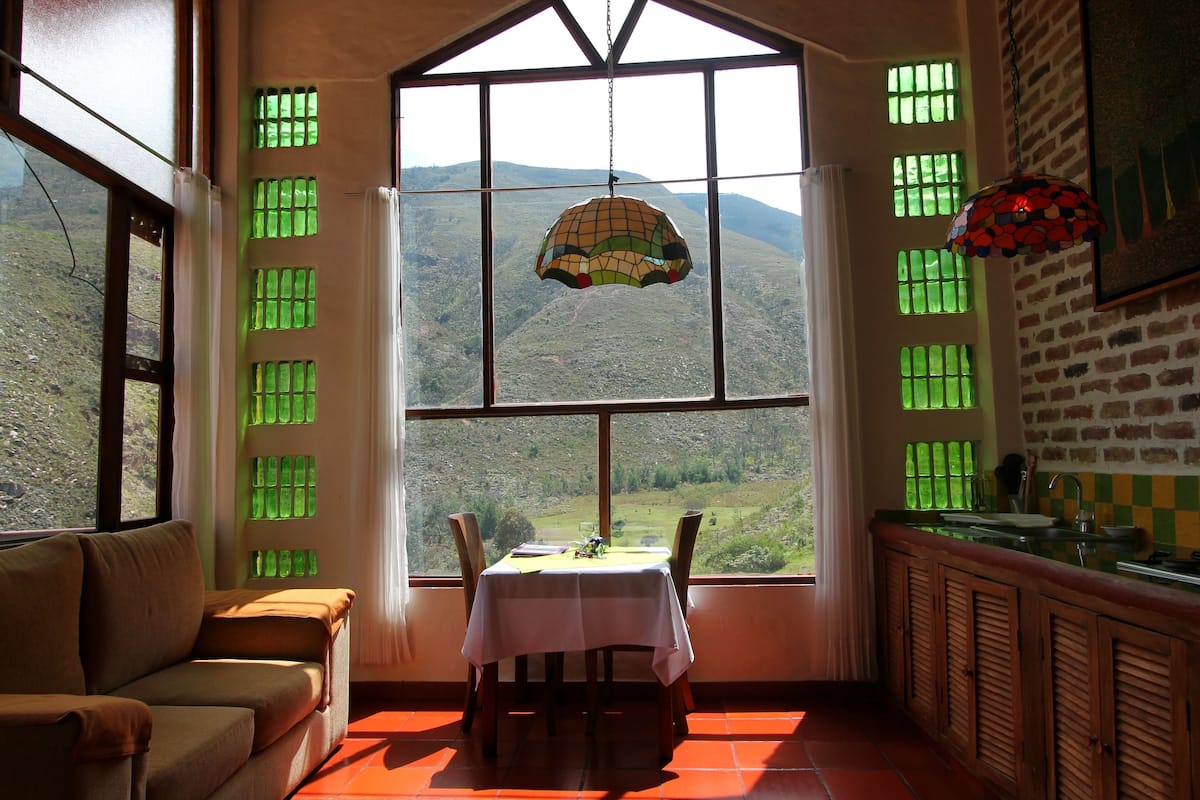 Un comedor con una mesa preparada para dos personas, un sofá y vistas a un paisaje montañoso a través de amplios ventanales.