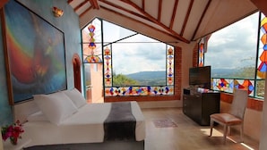 A bedroom with a bed, a TV, a desk, and large windows with a view. - Suites Arcoiris (Villa de Leyva)