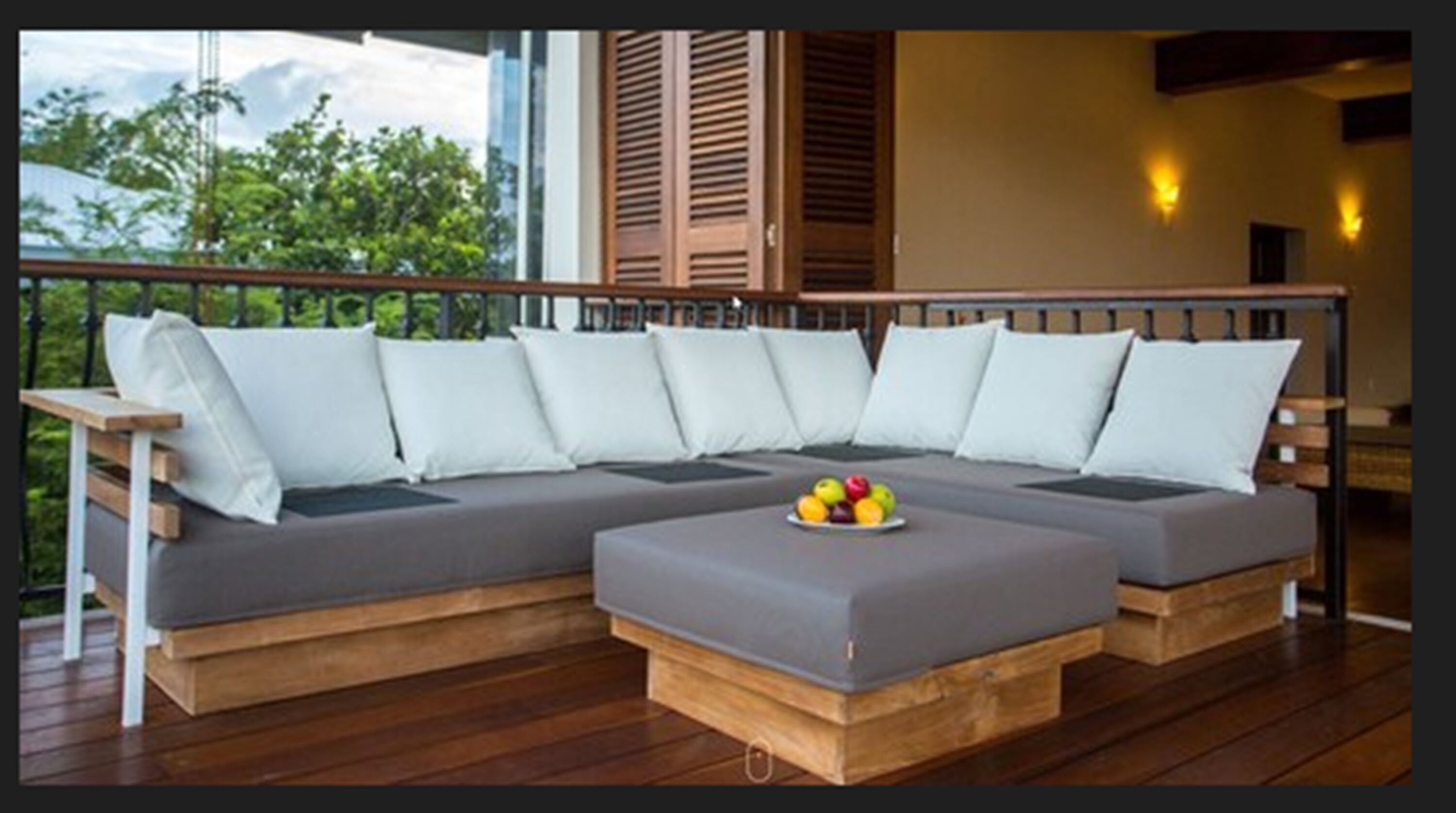 Un sofá modular con base de madera y cojines blancos, sobre una terraza de madera con barandilla.