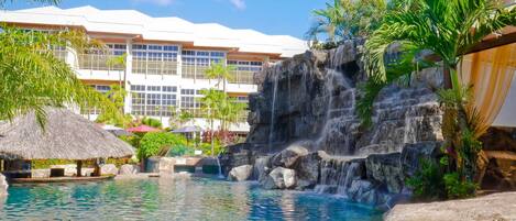 A resort pool with a waterfall, palm trees, and a building with balconies in the background.