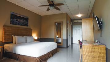 A hotel room with a large bed, a desk with a chair, a ceiling fan, a mounted TV, and a painting on the wall.