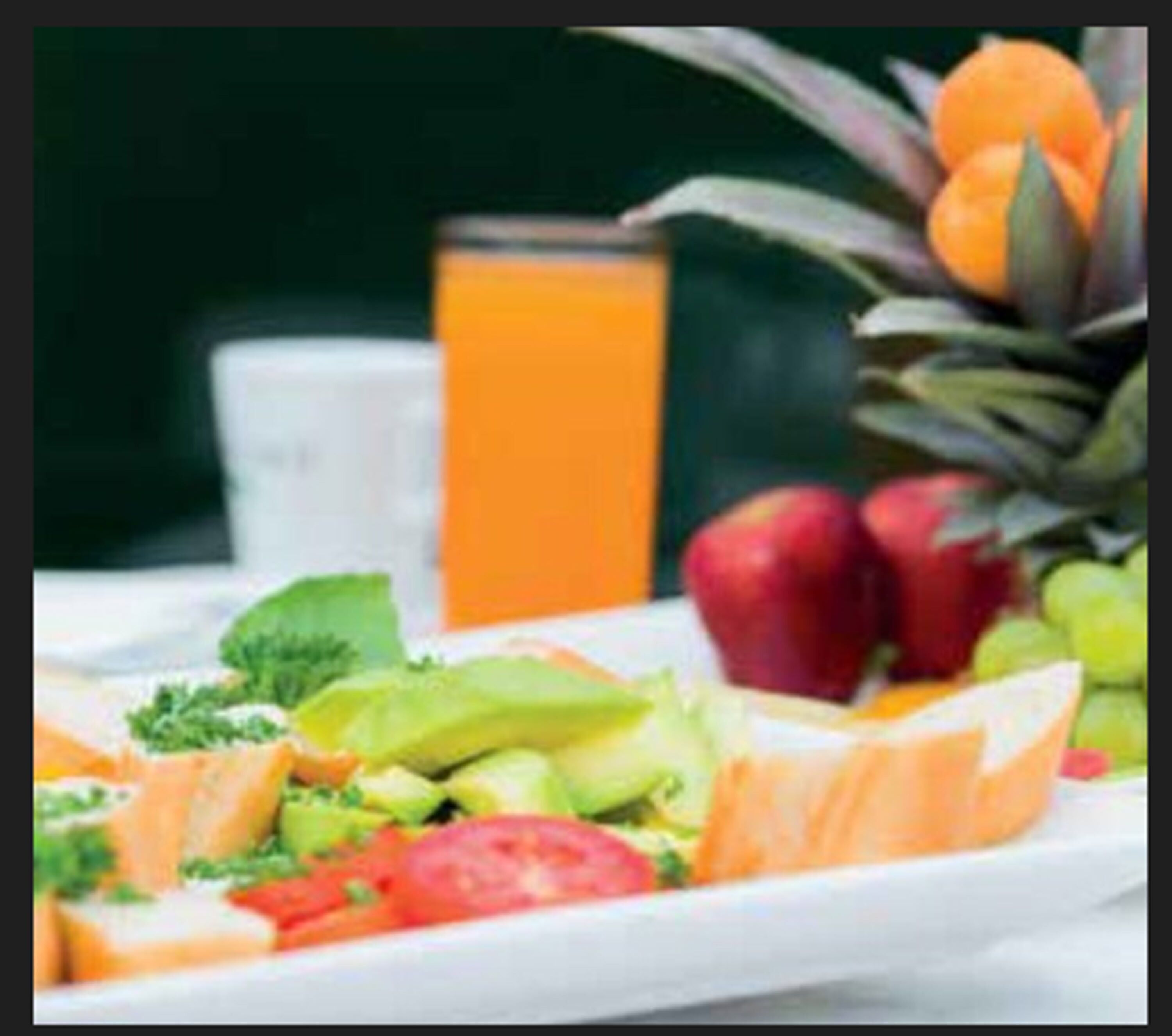 Ein Frühstücksteller mit Eiern, Avocado, Tomate und grünem Salat, ein Glas Orangensaft und eine Ananas mit Orangen im Hintergrund.