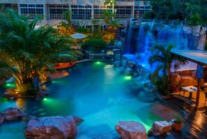 Piscina de resort con cascada, rodeada de plantas tropicales y palmeras.