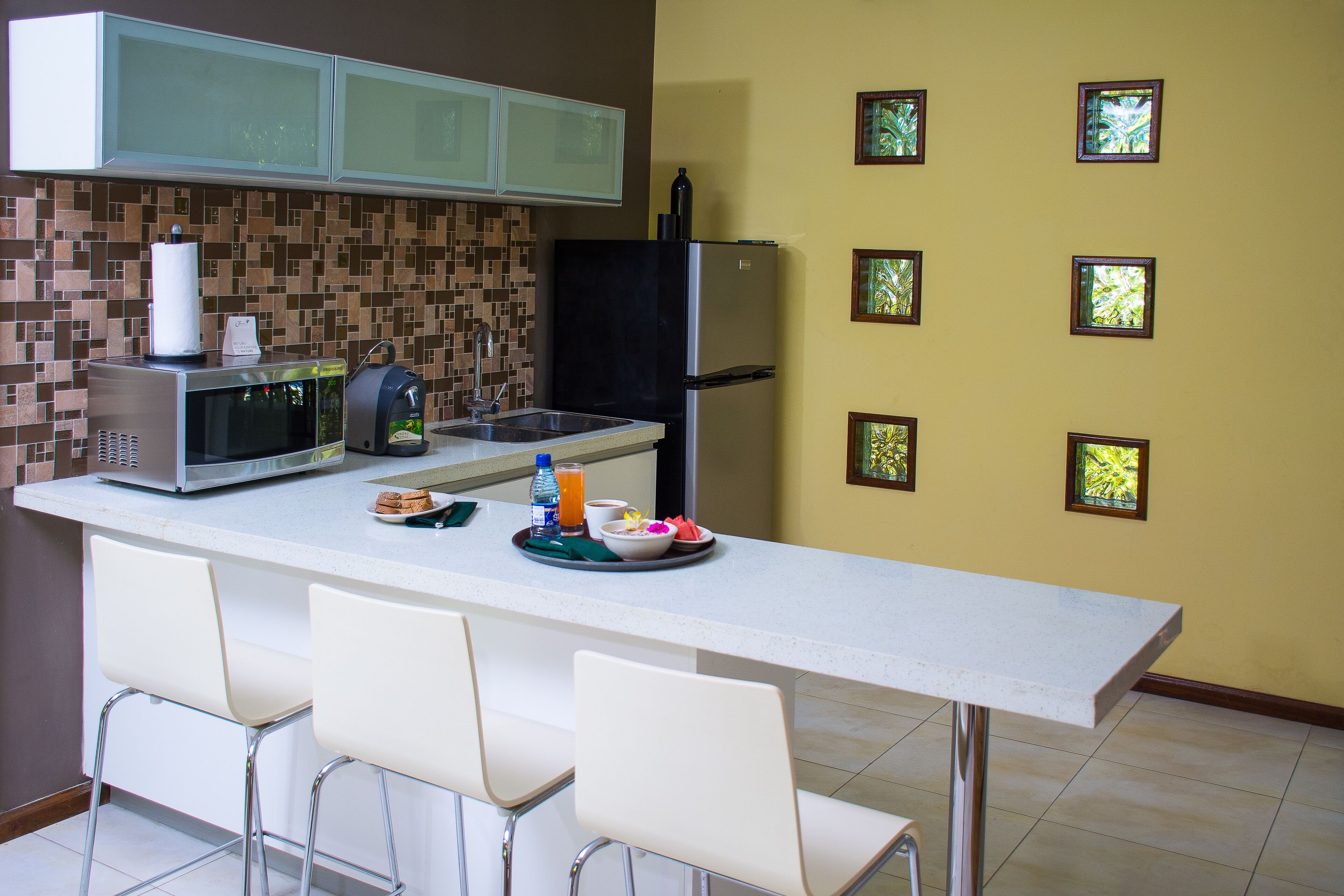 A kitchen with a white countertop, a refrigerator, a microwave, and a sink. There are four white chairs around the counter.