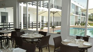 A dining area with tables and chairs, large windows, and a view of a swimming pool and outdoor area. - Ocean Blue Hotel (Manglaralto)