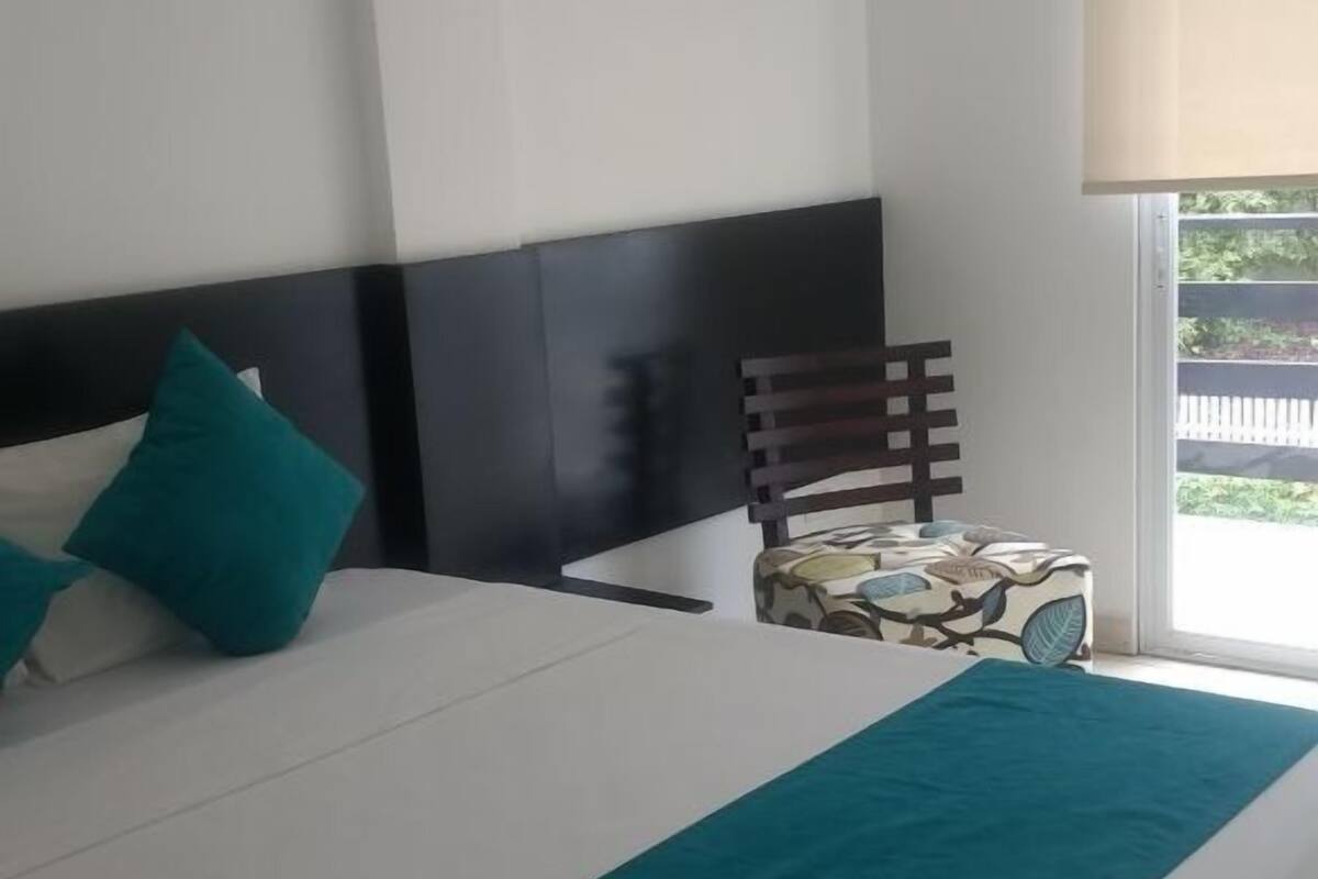 Habitación de hotel con una cama grande, una mesita de noche, una silla y una ventana con vista a follaje.