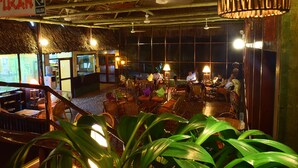 Un restaurant intérieur doté d’un toit de chaume, de meubles en osier et d’un éclairage d’ambiance.