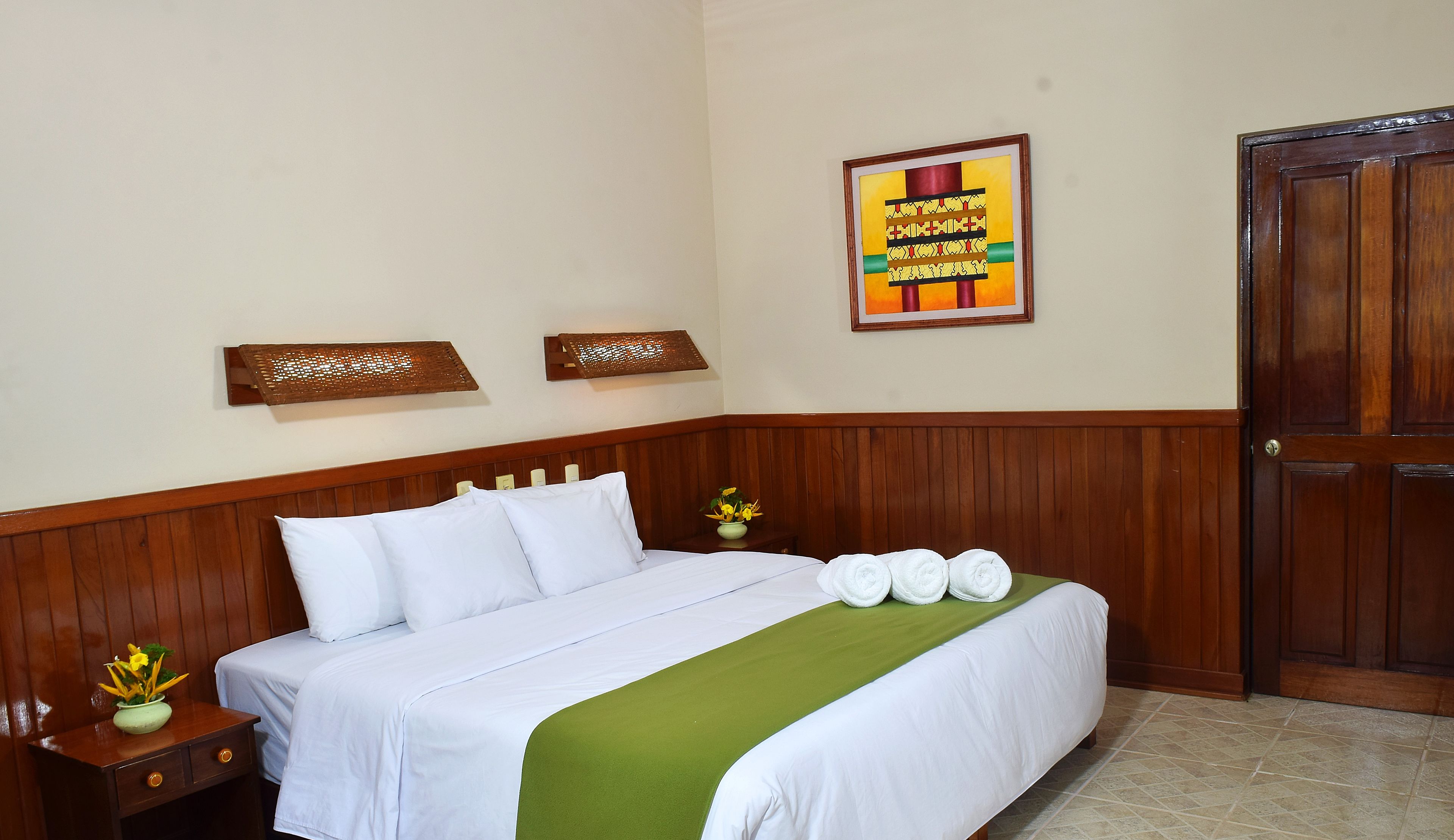 A neatly made bed with white linens, a green throw blanket, and a set of towels. There are two wall-mounted lamps and a framed picture above the bed.