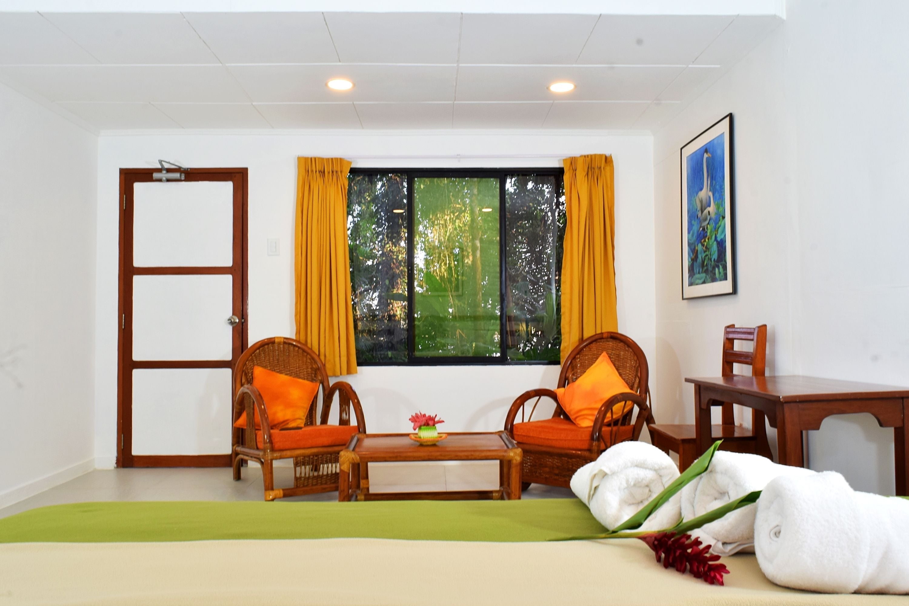 A room with two wicker chairs, a small table with a vase, a bed with a green bedspread, and a window with yellow curtains.