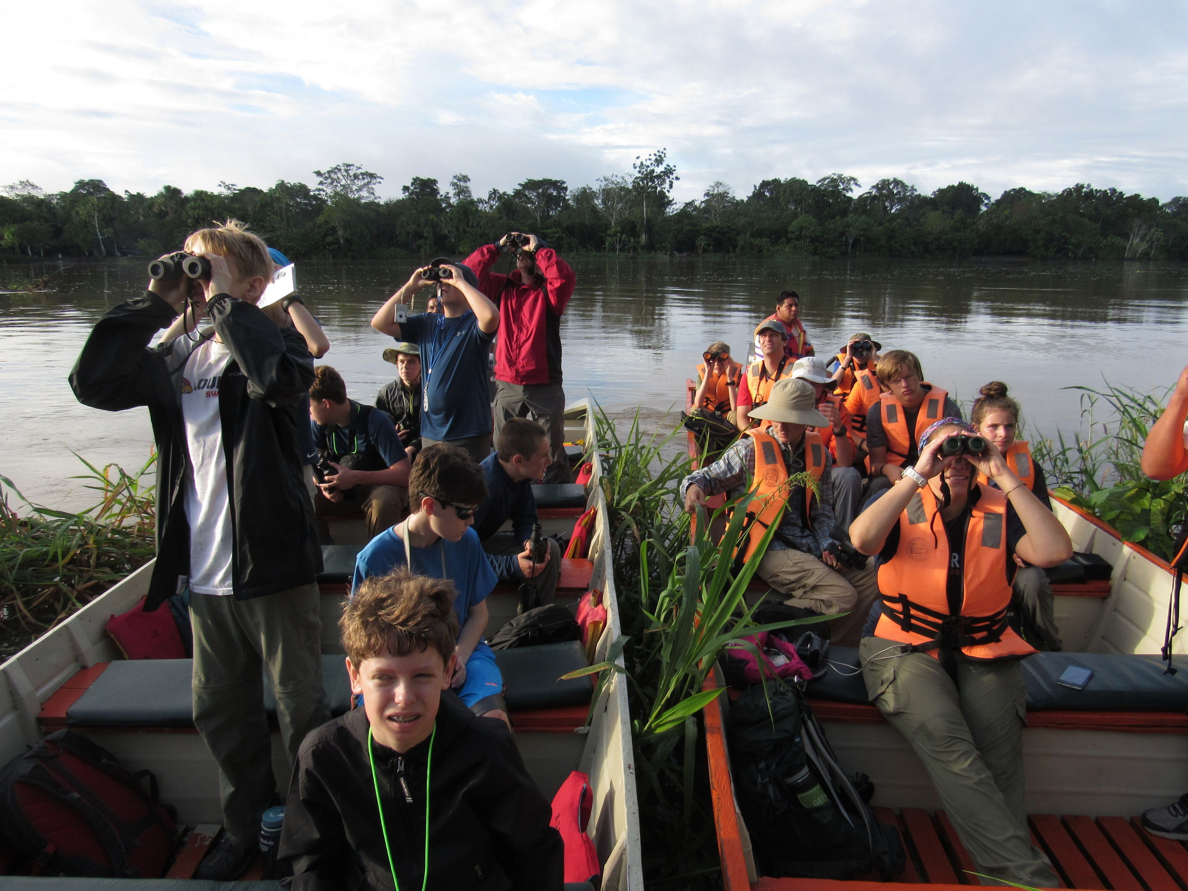 A group of people on a boat, some wearing life jackets, with a calm lake and trees in the background.
