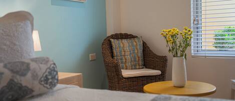 A bedroom with a bed, a wicker chair with a cushion, a small round table with a vase of flowers, and a window with blinds.