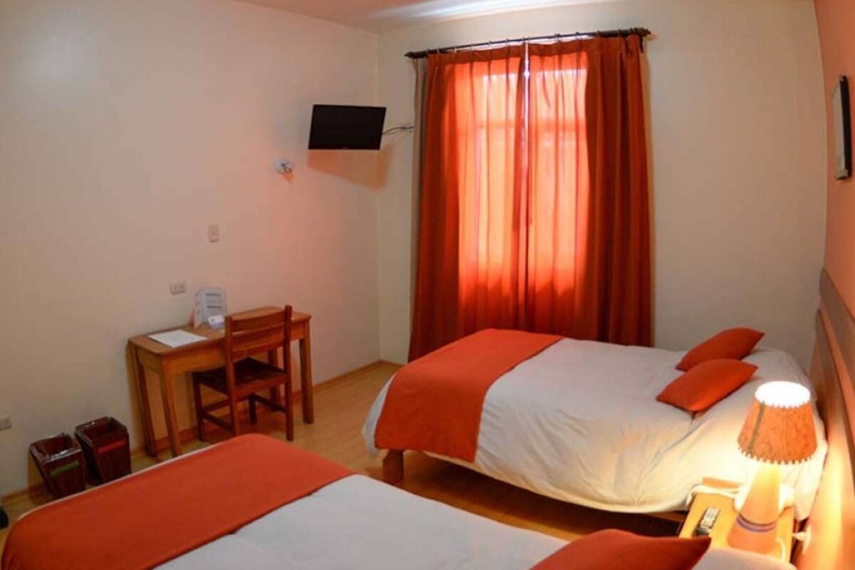 Ein Hotelzimmer mit zwei Betten, einem Schreibtisch, einem Sessel, einem an der Wand befestigten Fernseher und einem Fenster mit Vorhängen.