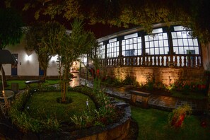 A well-lit garden area with a stone wall, a wooden bench, and a pathway leading to a building with large windows.