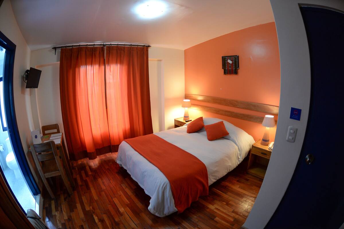 Ein Hotelzimmer mit Holzboden, einem Bett mit weißer und orangefarbener Bettwäsche, einem Nachttisch mit Lampe und einem an der Wand befestigten Fernseher.