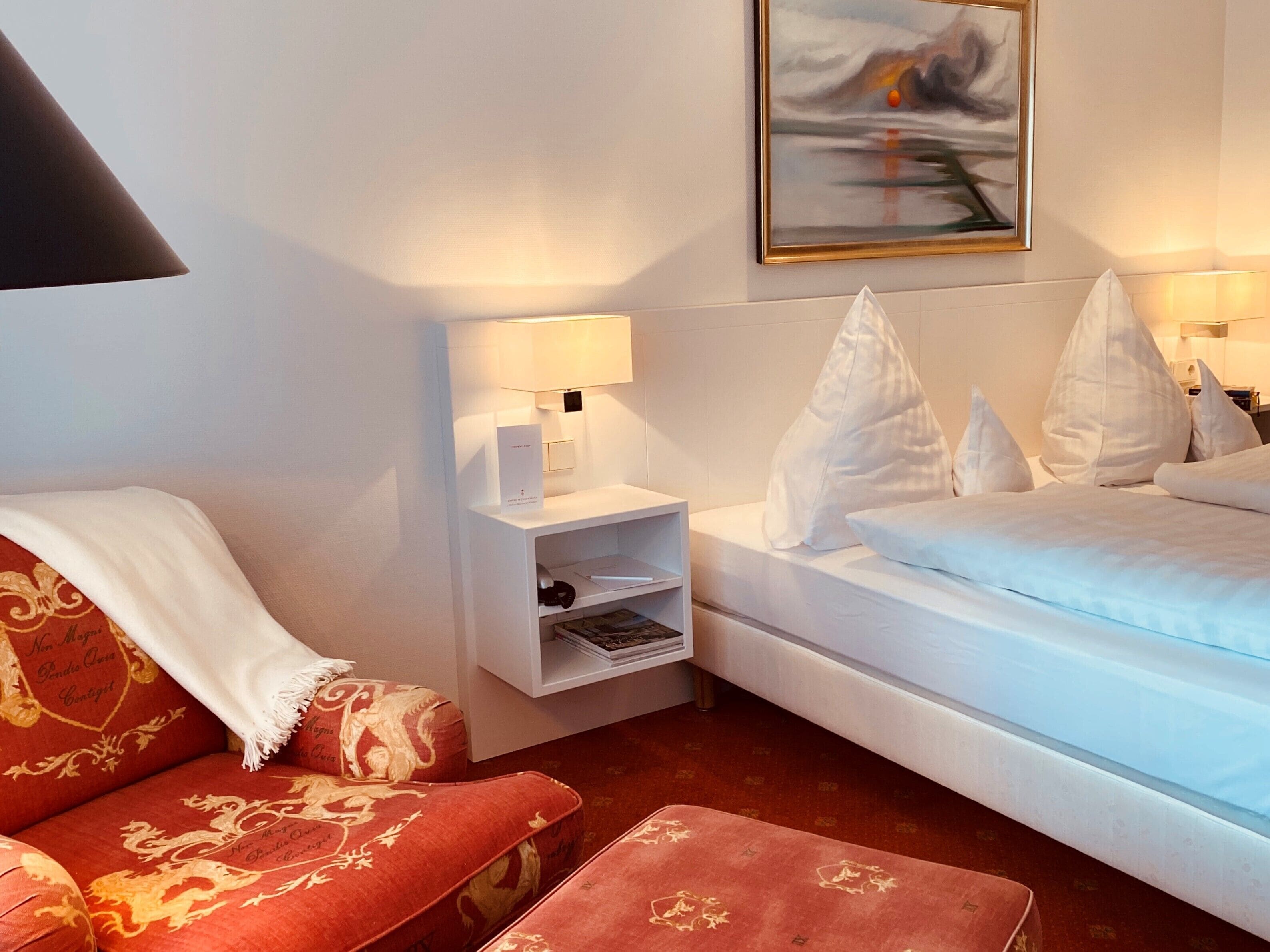 A hotel room with a bed, a red upholstered sofa, a white nightstand with a lamp, and a painting on the wall.