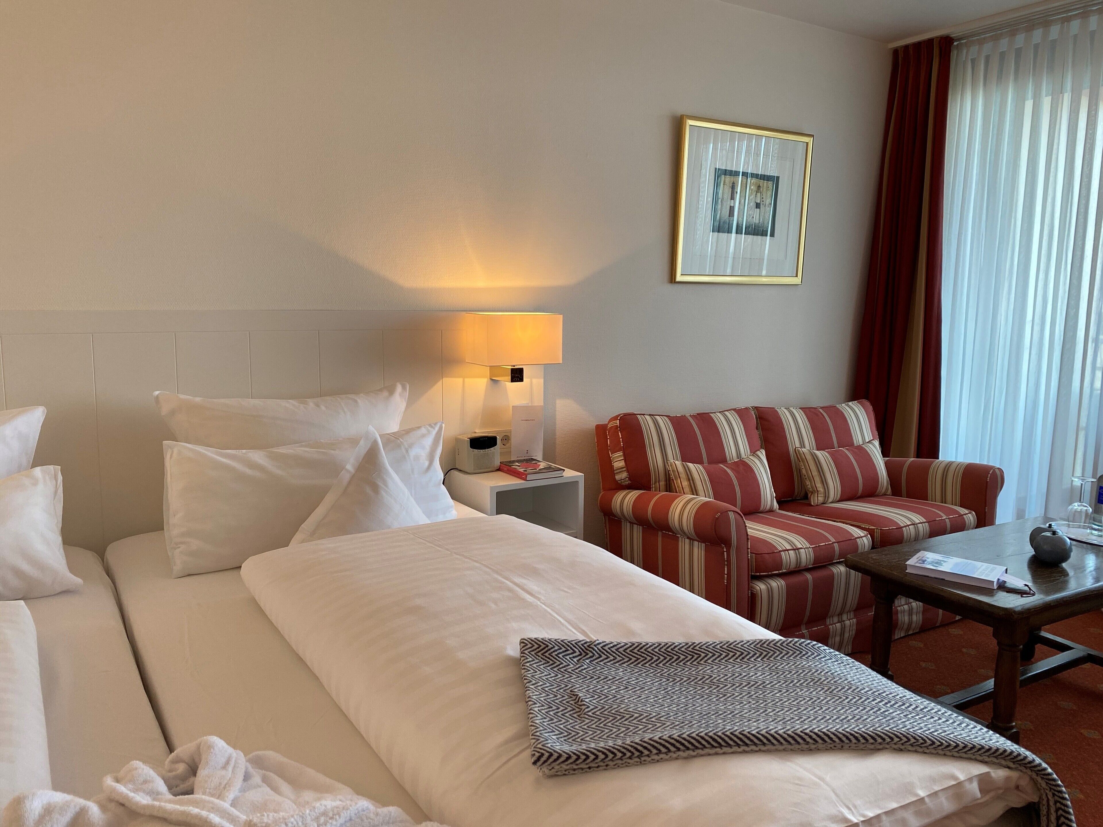 Habitación de hotel con cama, sofá, una mesita, lámpara, un cuadro en la pared y ventana con cortinas.