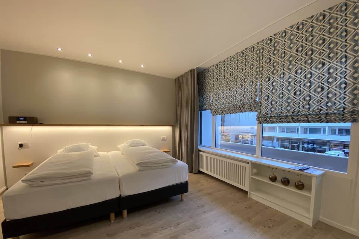 A modern hotel room with a large bed, a bedside table, a built-in shelf with decorative items, and a window with a patterned curtain.