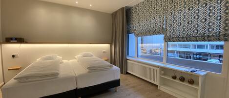 A modern hotel room with a large bed, a bedside table, a built-in shelf with decorative items, and a window with a patterned curtain.
