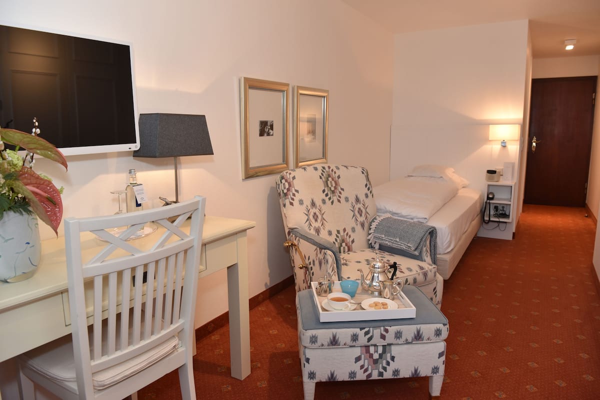 A hotel room with a desk, a chair, a bed, a bedside table, a lamp, a TV, framed pictures, and a small table with a tray of breakfast items.