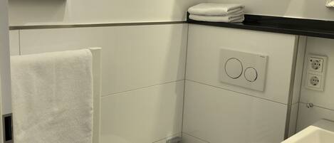 A small, modern bathroom with a toilet, a towel rack, a glass shelf with toiletries, and a sink.
