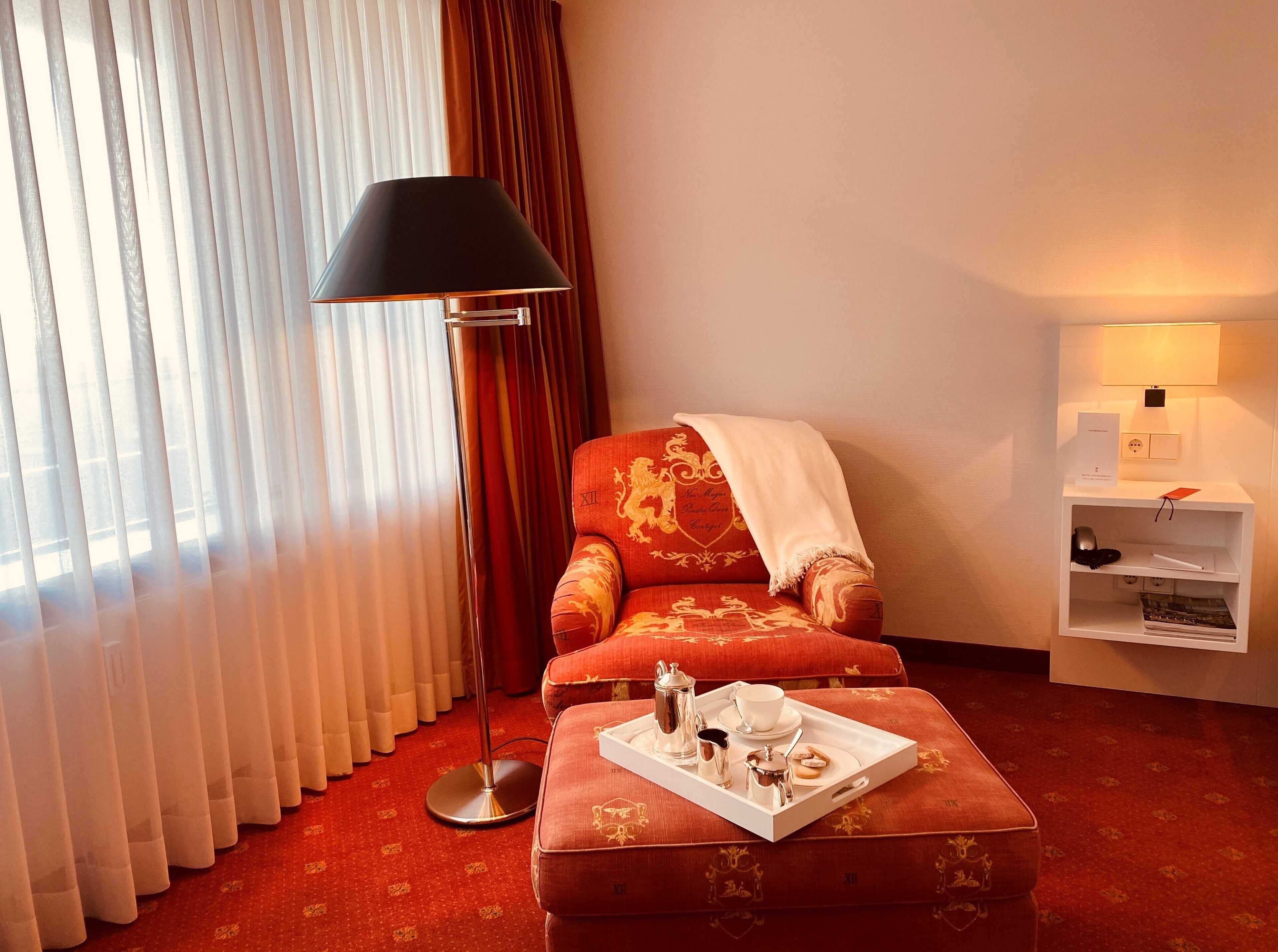 A hotel room with a red armchair, a white ottoman with a tray of tea and cream, a floor lamp, and a side table with a lamp and a sign.