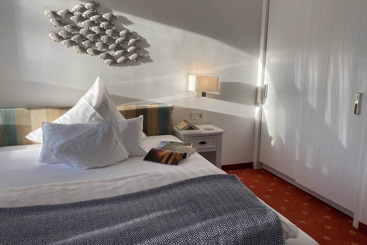 A hotel room with a bed, pillows, a bedside table with a lamp, a magazine, and a wall decoration of fish.