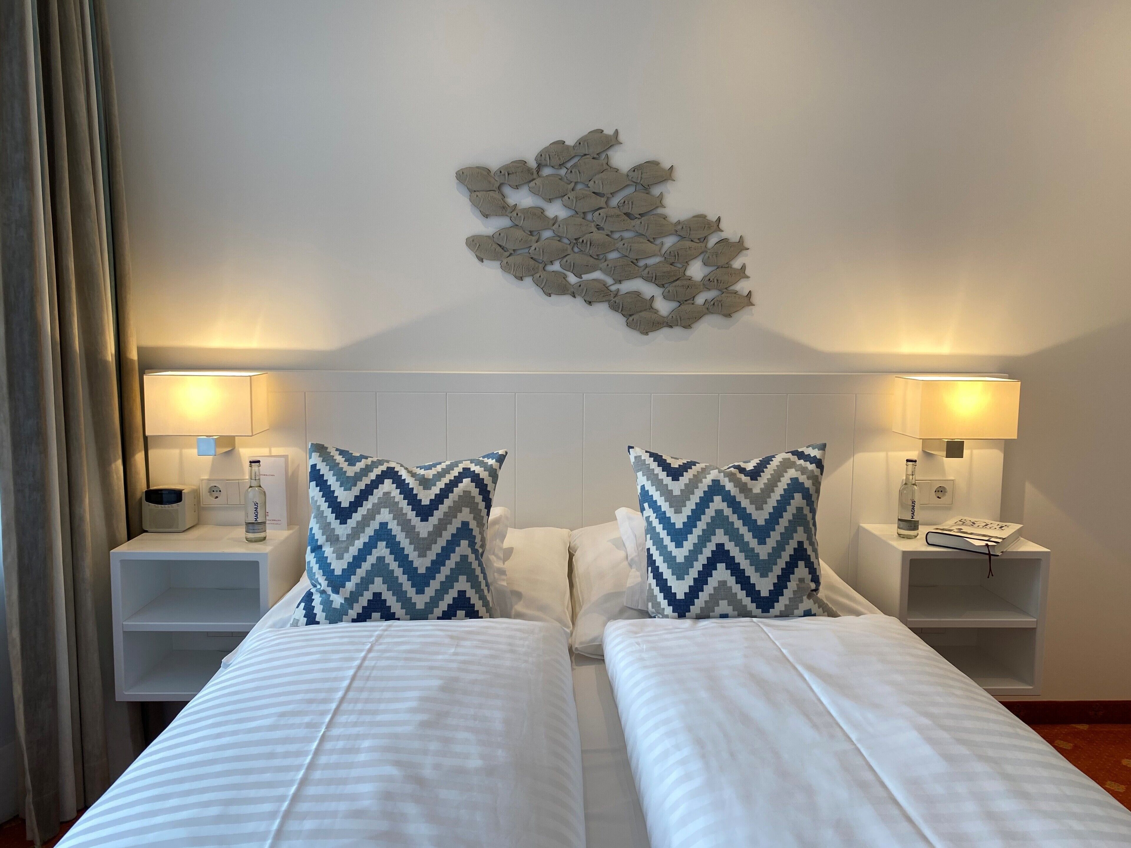 A hotel room with two beds, white bedding, a decorative wall piece, bedside lamps, and a nightstand with a bottle and book.