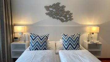 Camera d'albergo con due letti, biancheria da letto bianca, un elemento decorativo a parete, lampade sui comodini e un comodino con una bottiglia e un libro.