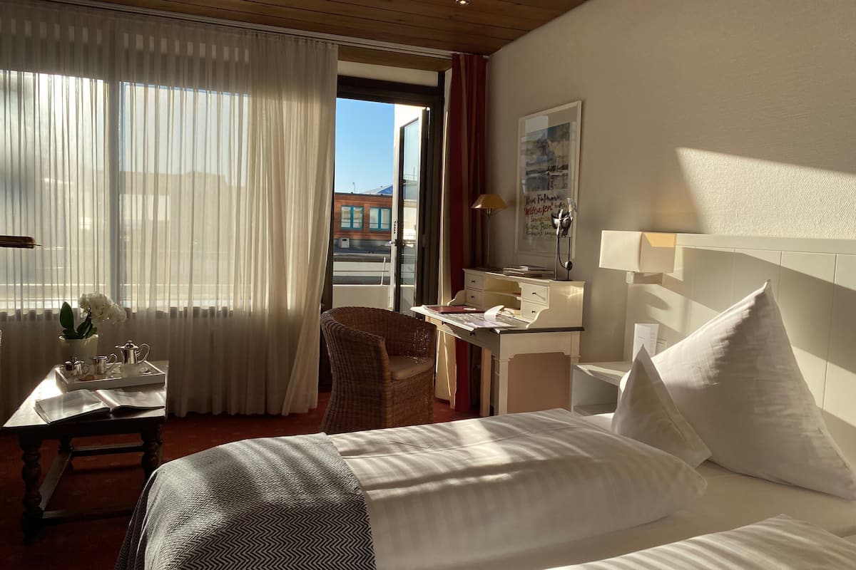 A hotel room with a bed, a desk with a chair, a window with sheer curtains, and a view of the outside.