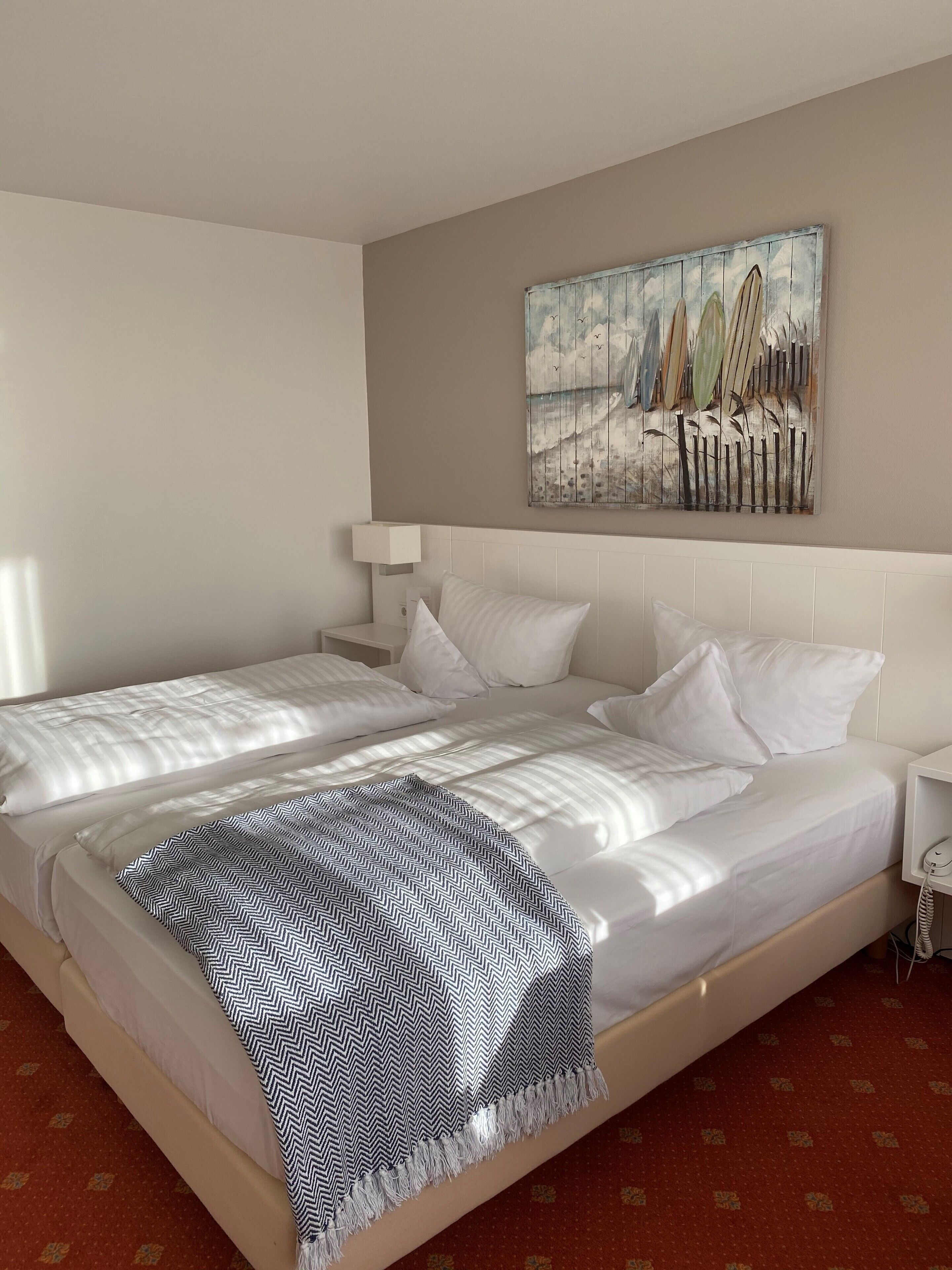Ein Hotelzimmer mit einem großen Bett, weißer Bettwäsche, einer gemusterten Tagesdecke, einem Nachttisch mit einer Lampe und einem Bild an der Wand.