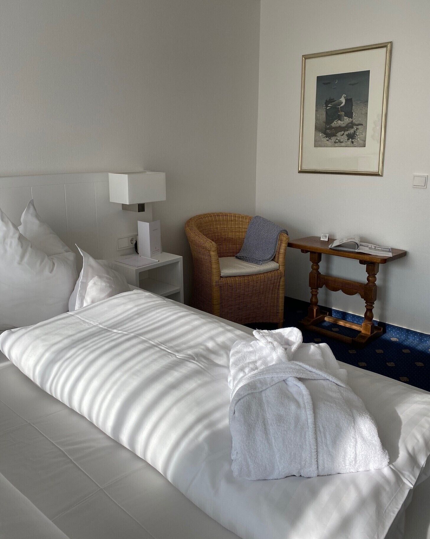 Camera d'albergo con un letto grande, una poltrona in vimini, un comodino e un quadro incorniciato appeso al muro.