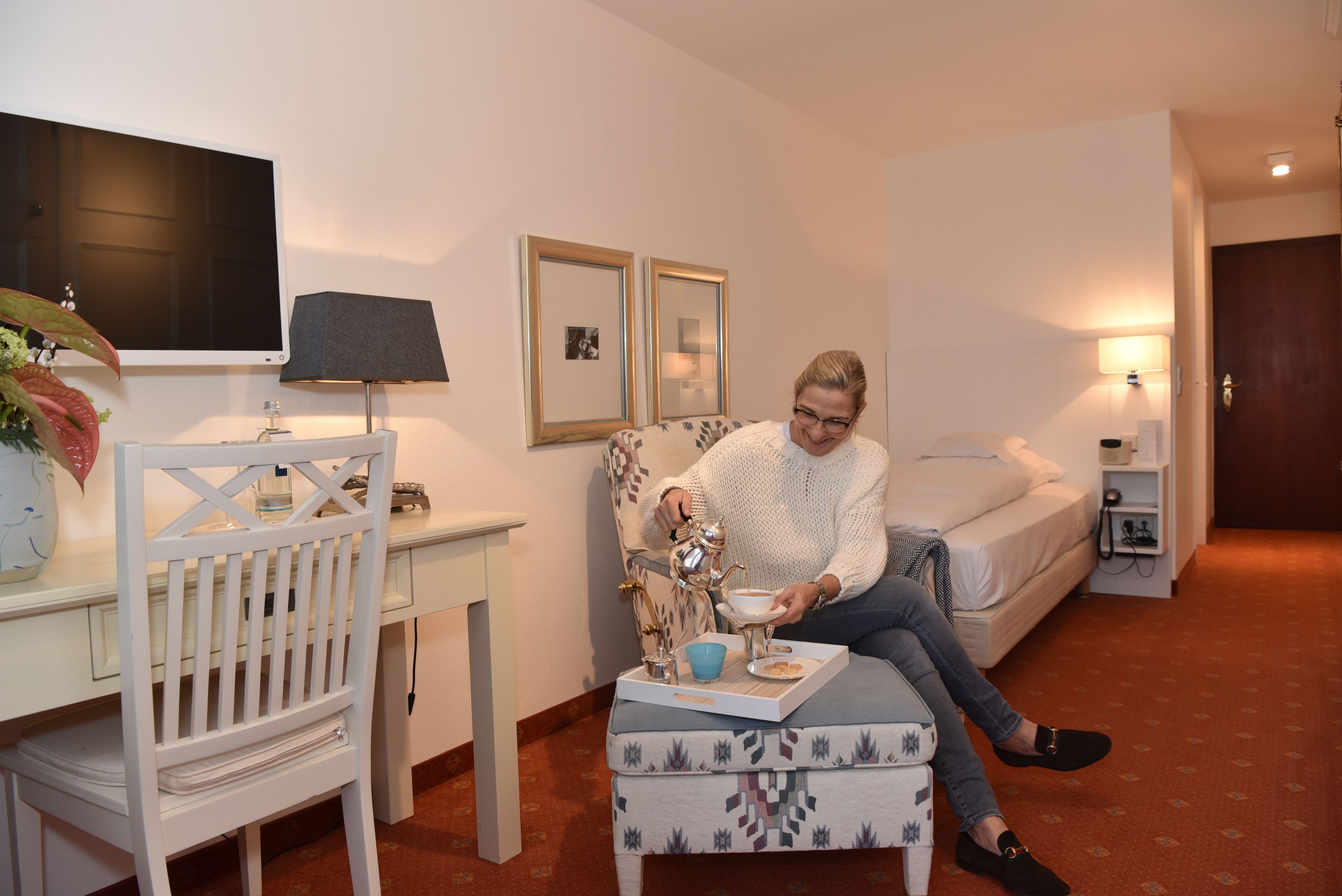 A hotel room with a bed, a desk with a chair, a small table with a tray of tea and biscuits, and a TV mounted on the wall.