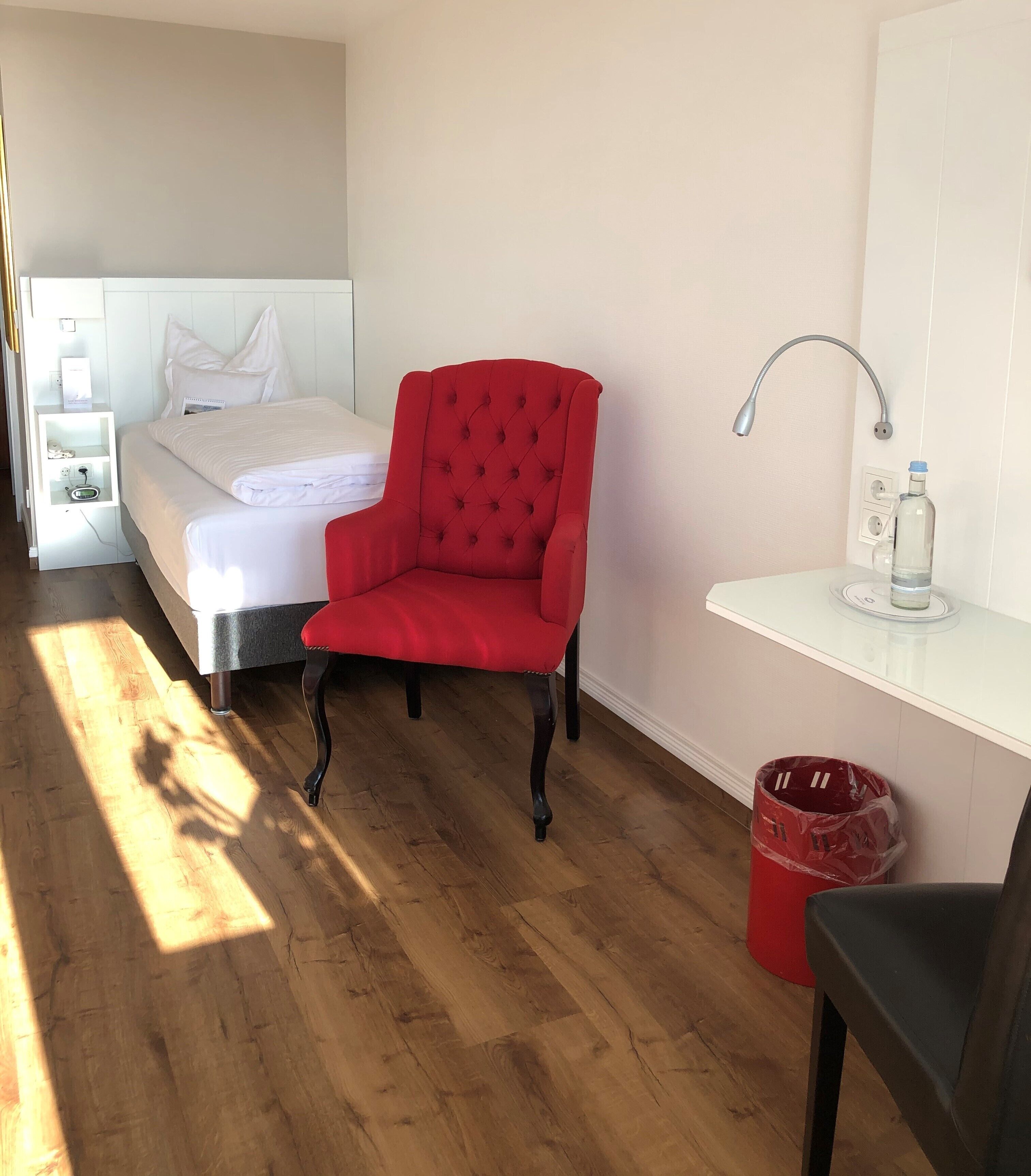 A hotel room with a bed, a red armchair, a desk with a lamp, and a trash bin.