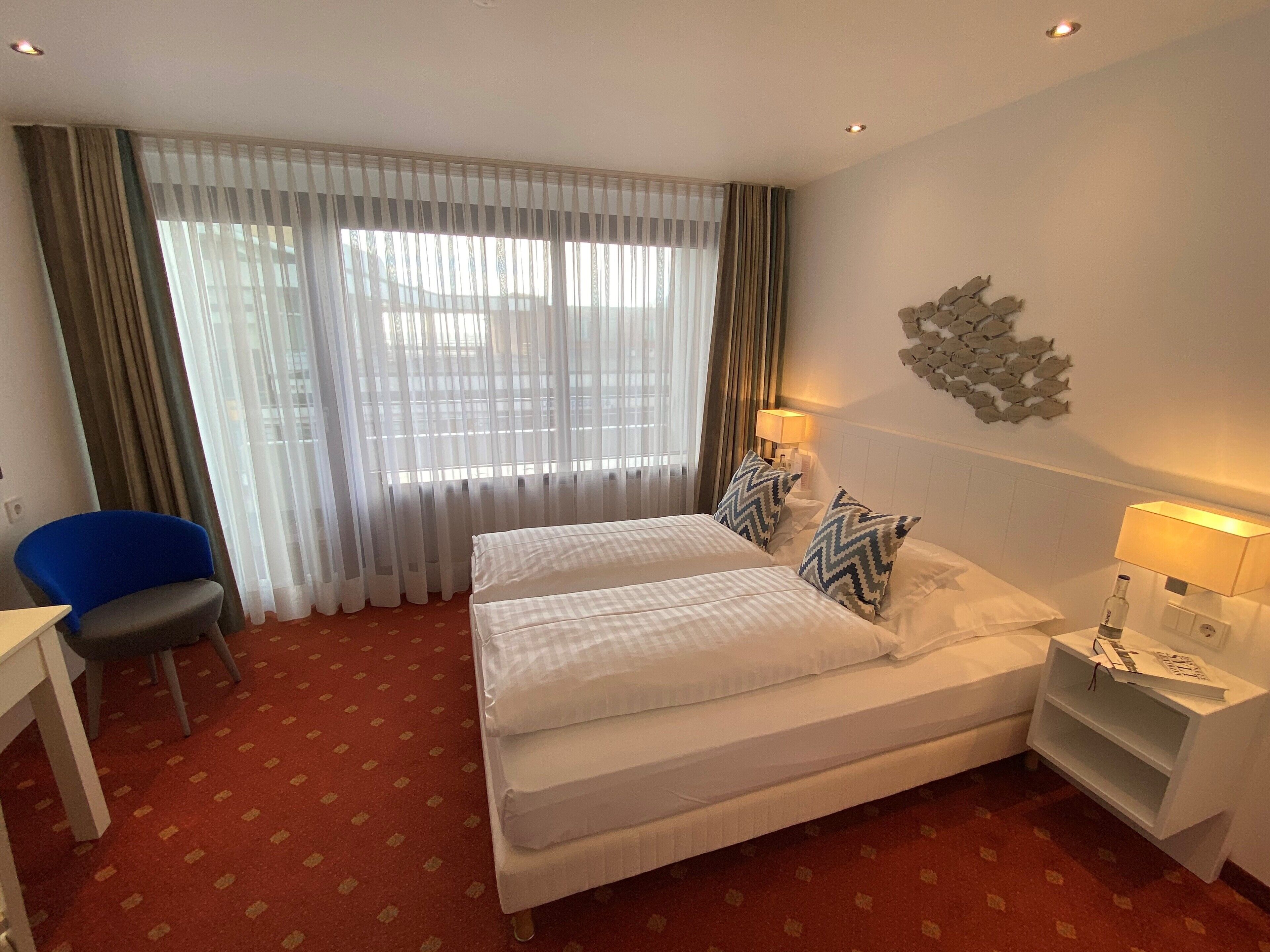 A hotel room with a double bed, two bedside tables with lamps, a chair, a desk, and a large window with curtains.