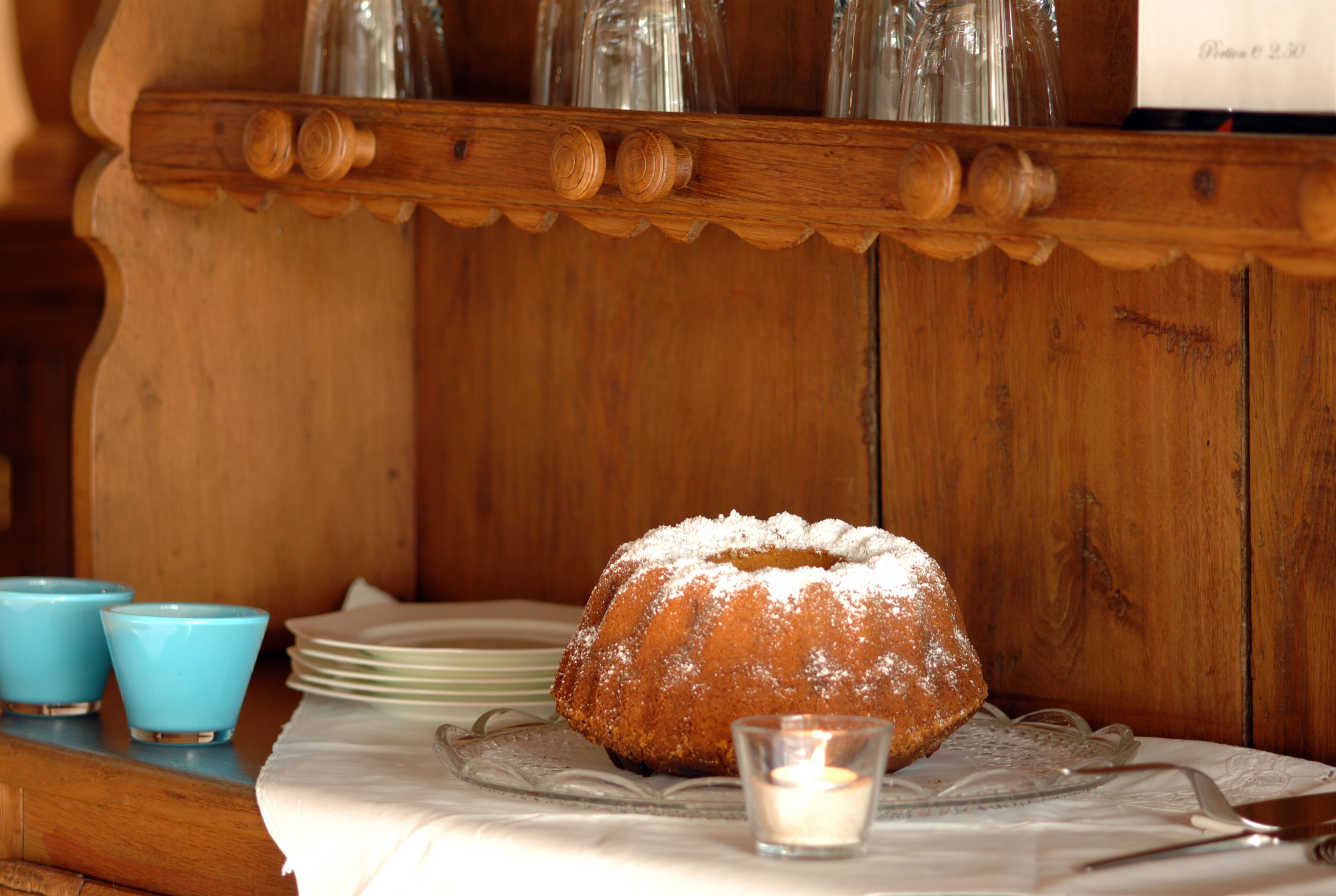 Een houten tafel met een bundt cake bestrooid met poedersuiker, een brandende kaars en blauwe bekers.