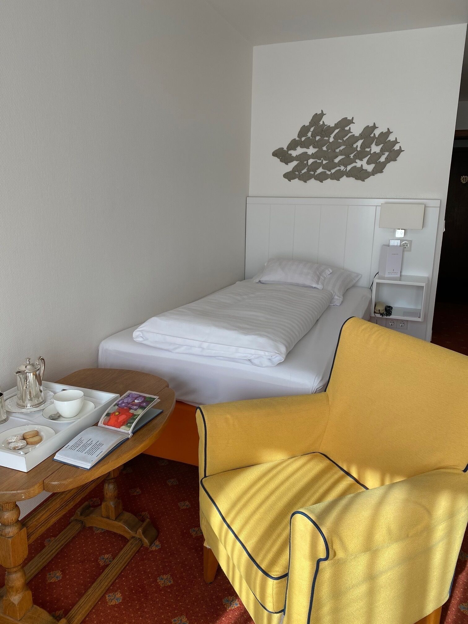 A hotel room with a single bed, a bedside table with a lamp, a small table with a tray of breakfast items, and a yellow armchair.