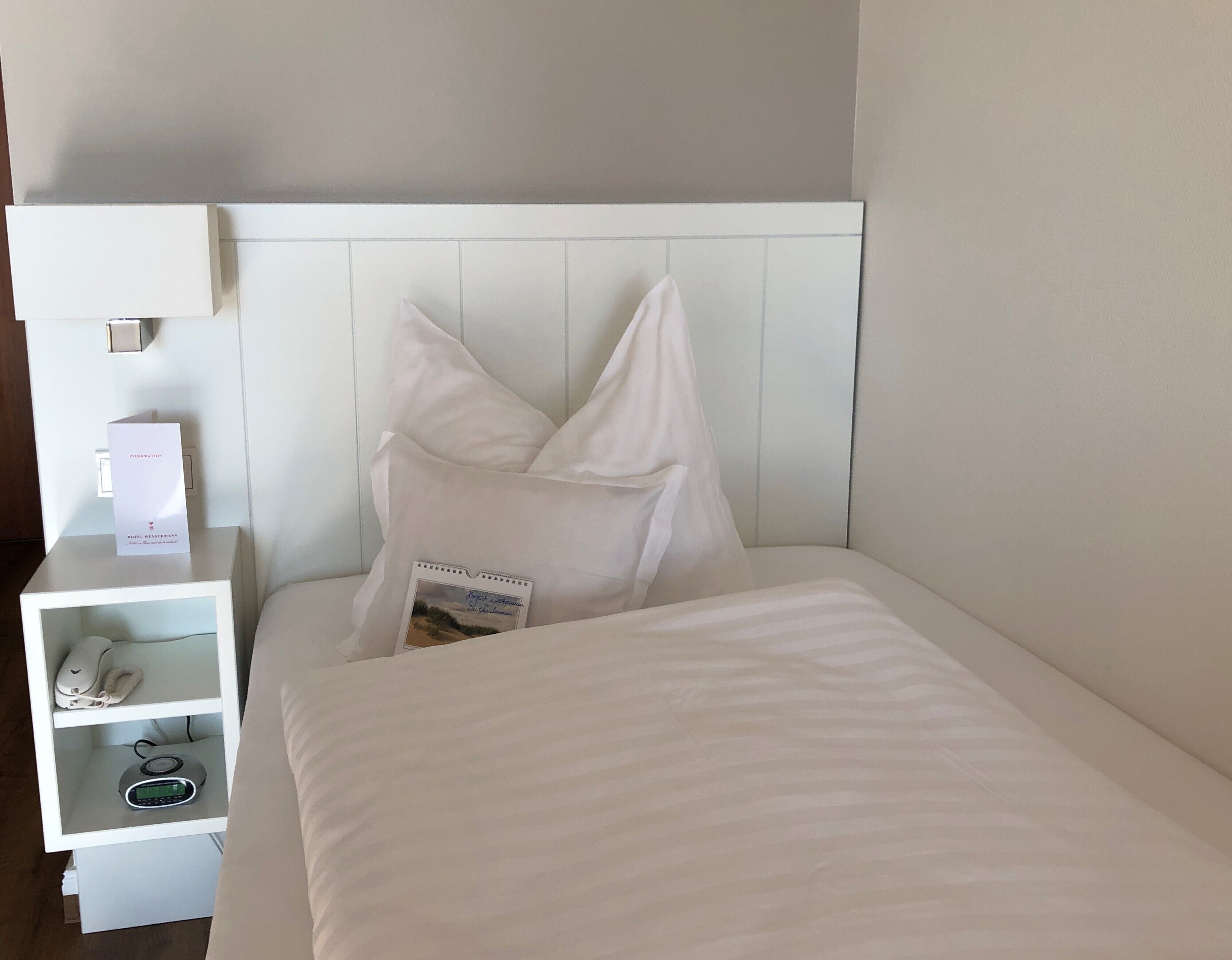 A neatly made bed with white bedding, a white nightstand with a phone and a digital device, and a wall-mounted light fixture.