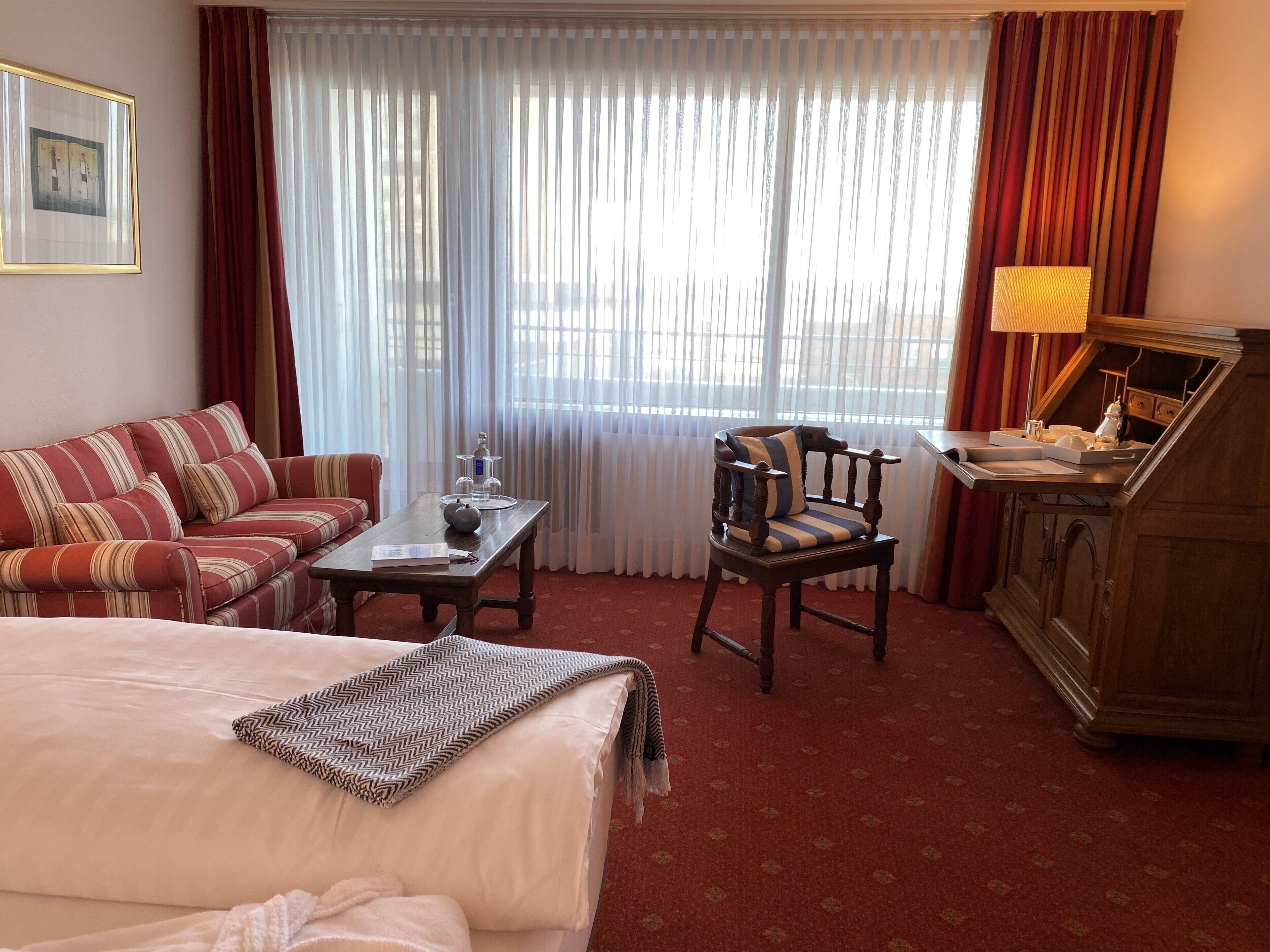 Hotellihuone, jossa on sänky, sohva, puinen tuoli, työpöytä lampun kanssa ja ikkuna, jossa on verhot.