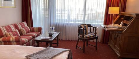 A hotel room with a bed, a sofa, a wooden chair, a desk with a lamp, and a window with curtains.