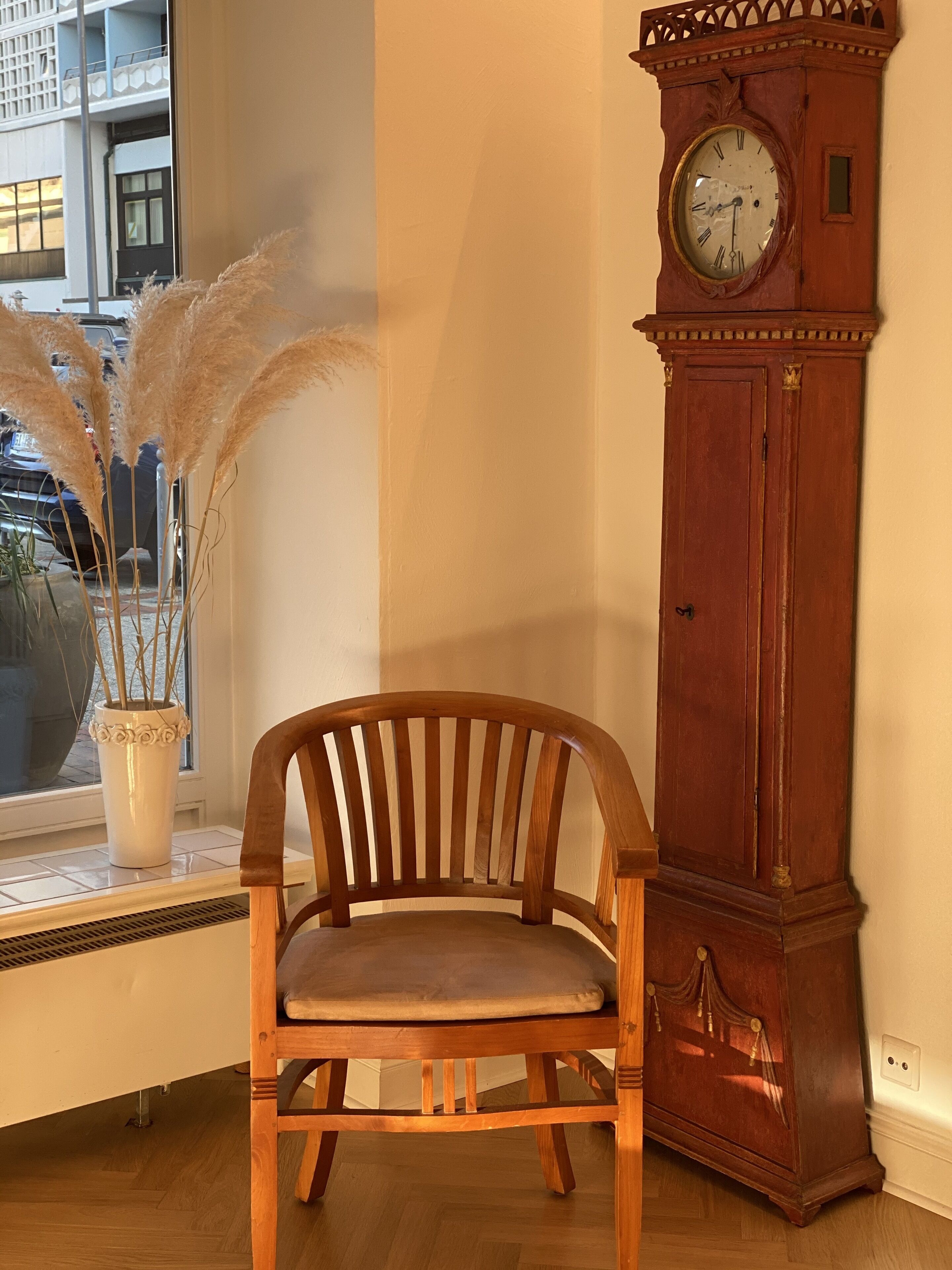 A wooden chair with a cushioned seat, a tall wooden clock, and a vase with pampas grass on a windowsill.