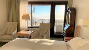 A hotel room with a bed, two armchairs, a small table with a lamp, a wooden cabinet, and a view of the city through a large window. - Hotel Wünschmann (Sylt)