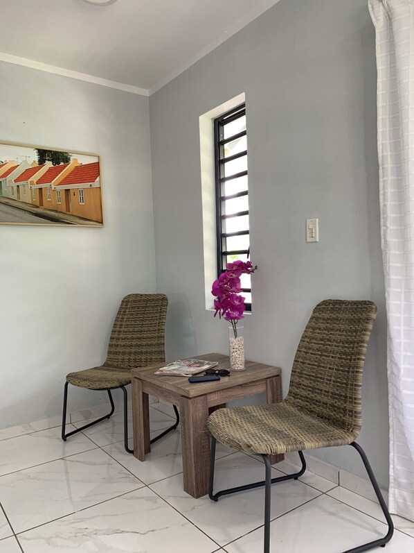 A room with two wicker chairs, a small wooden table, a vase with purple flowers, and a window with blinds. - Lilu Apartments Curacao (Willemstad)
