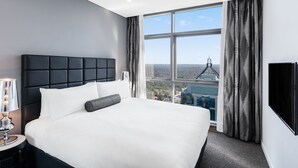 Una habitación de hotel moderna con una cama grande, una mesita de noche, un televisor y un ventanal con vistas a la ciudad.