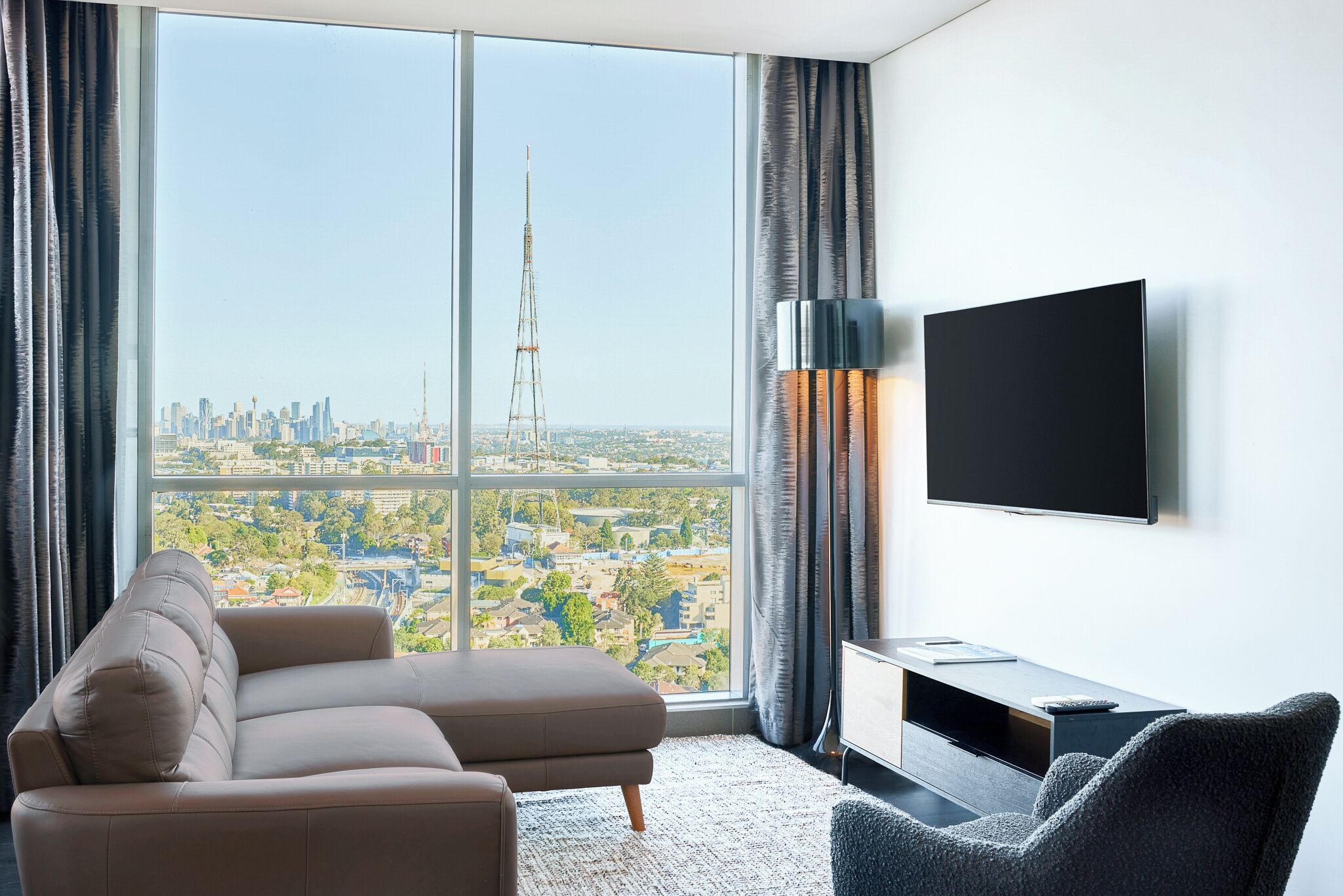 A modern hotel room with a large window offering a city view, a flat-screen TV mounted on the wall, and a comfortable seating area with a sofa and a chair.