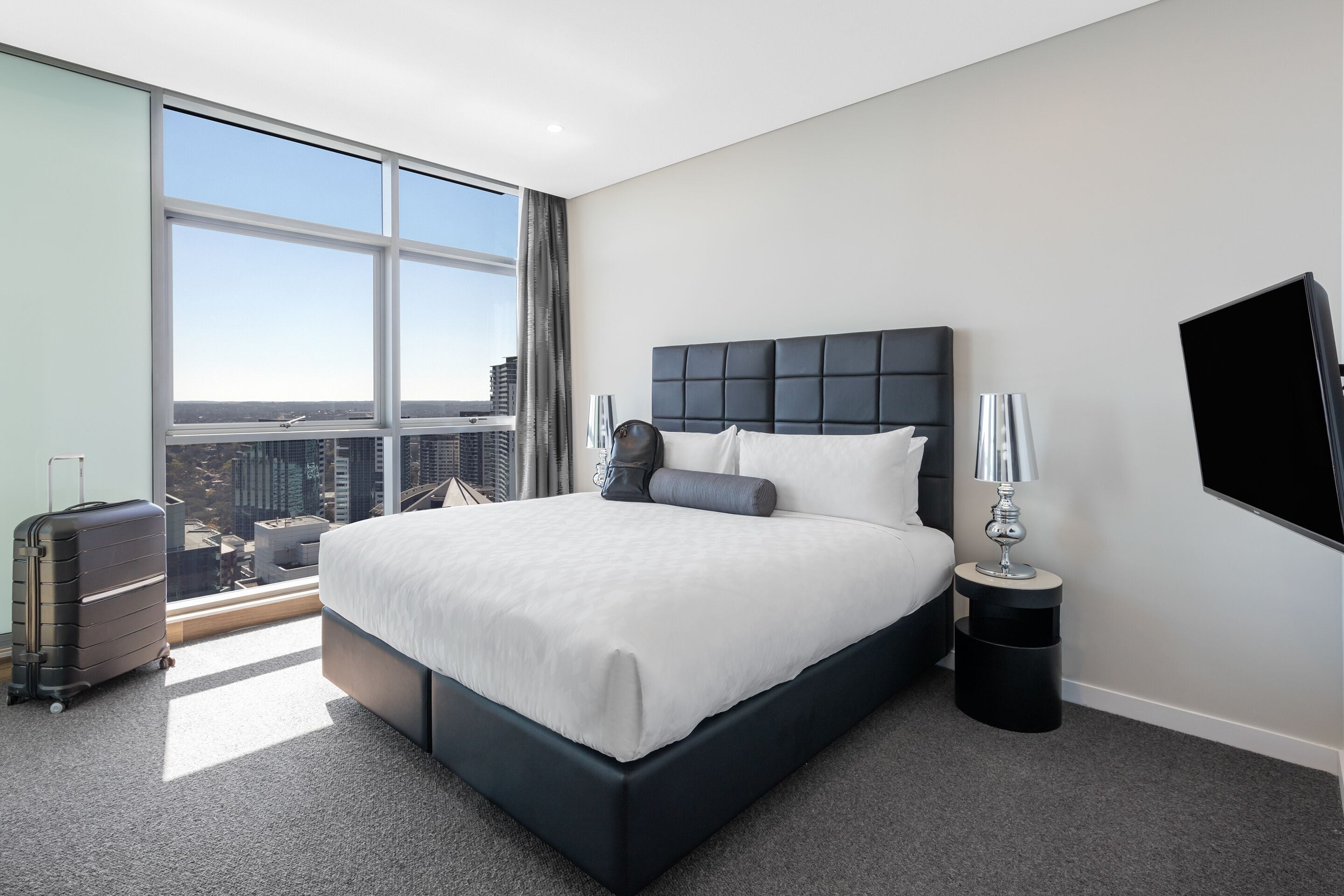 A modern hotel room with a large bed, a view of the city, a suitcase, and a TV on the wall.