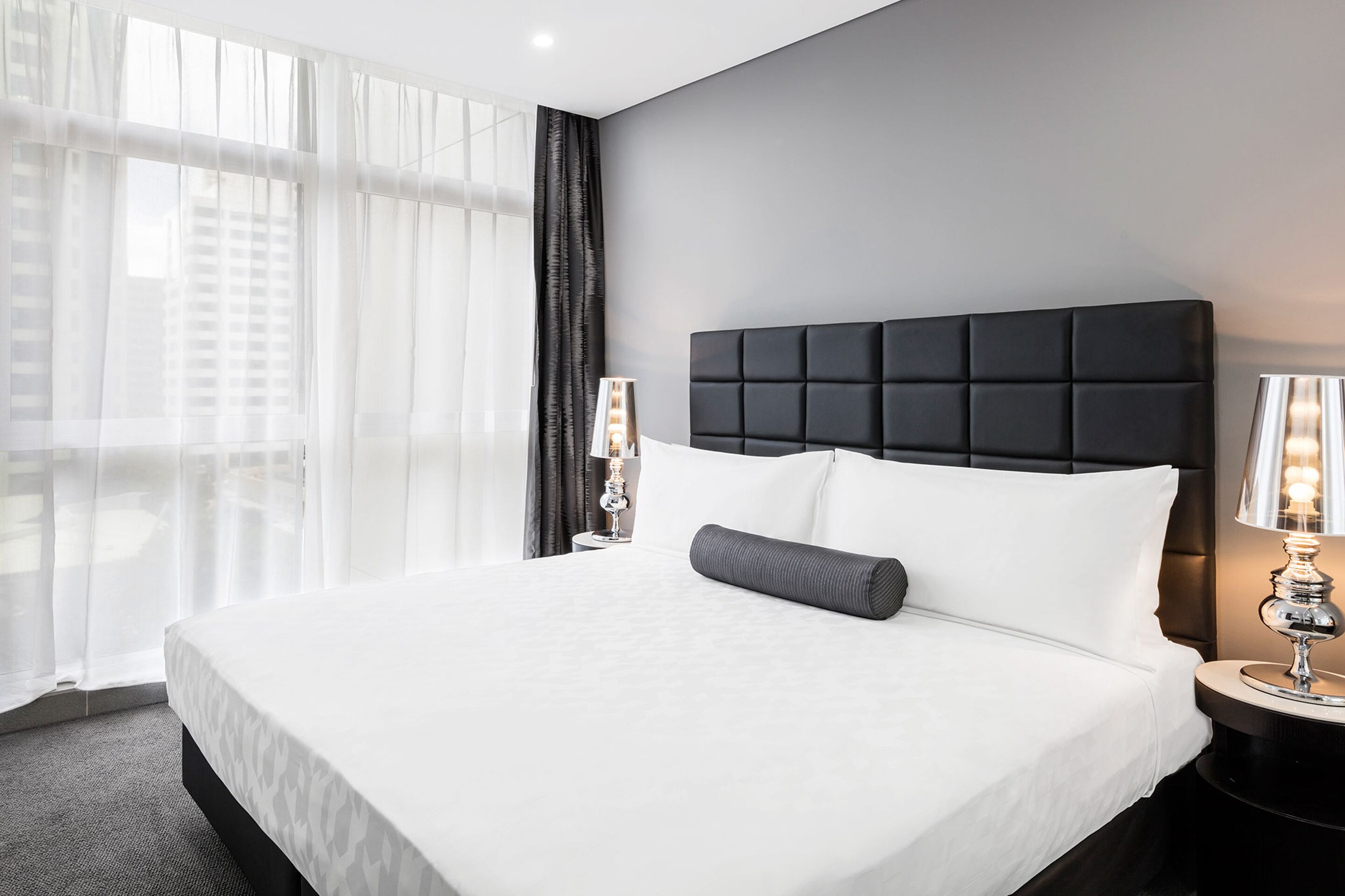 A hotel room with a large bed, two bedside lamps, and sheer curtains.