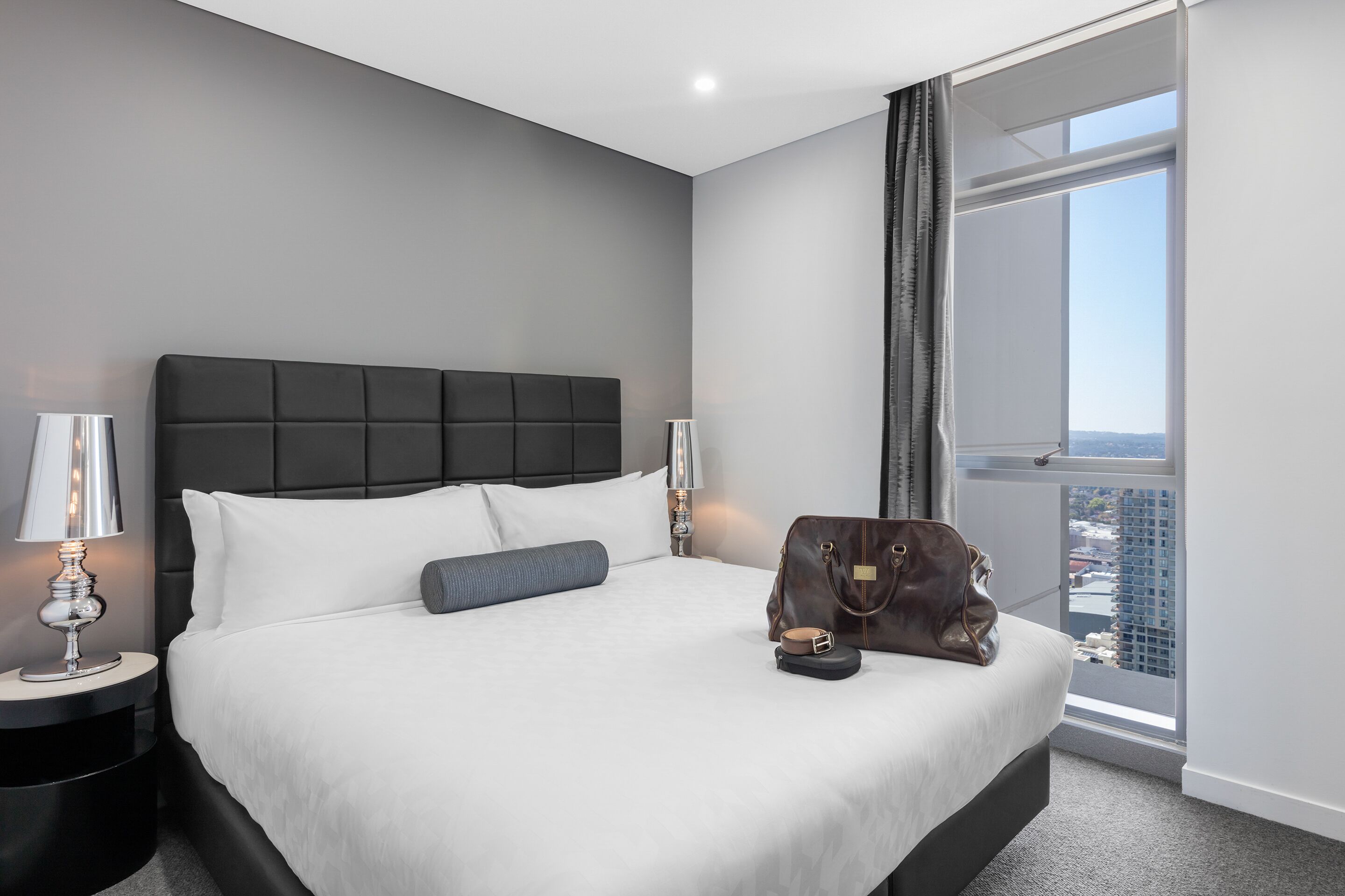 Een moderne hotelkamer met een groot bed, een nachtkastje met een lamp, en een stadszicht door een groot raam, plus een handtas op het bed.